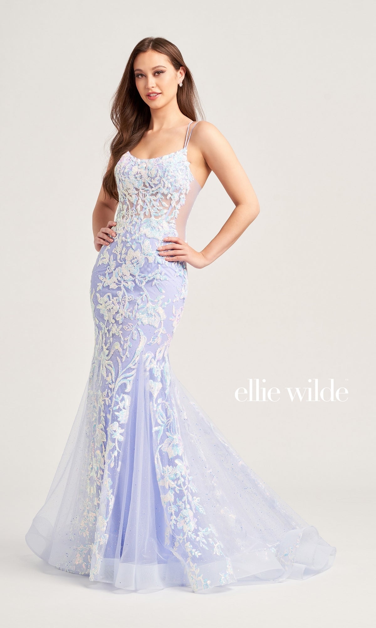A woman poses hands on hips in the Ellie Wilde Sequin Long Mermaid Prom Dress EW35008—an elegant fitted, pale lavender mermaid gown with floral lace appliqué and thin straps.